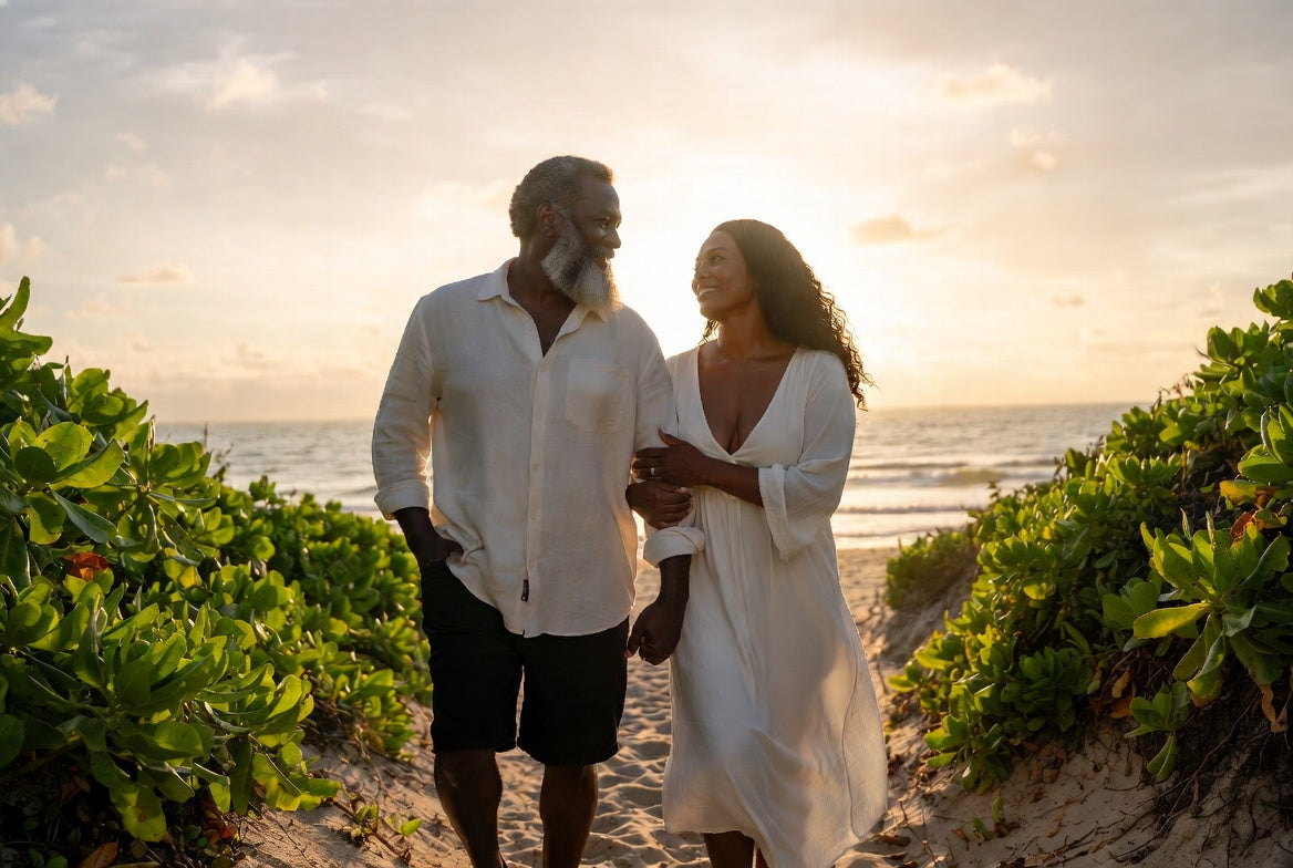A grey-bearded man and a woman walking a sandy path through coastal greenery at sunset, smiling at each other.