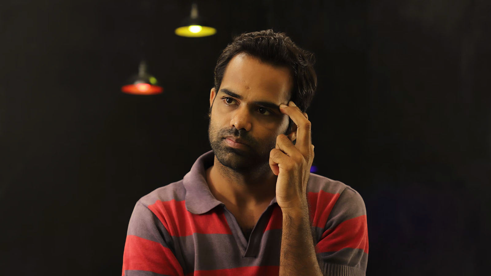 A man in a striped shirt resting a hand against his temple, looking thoughtful against a dark background.