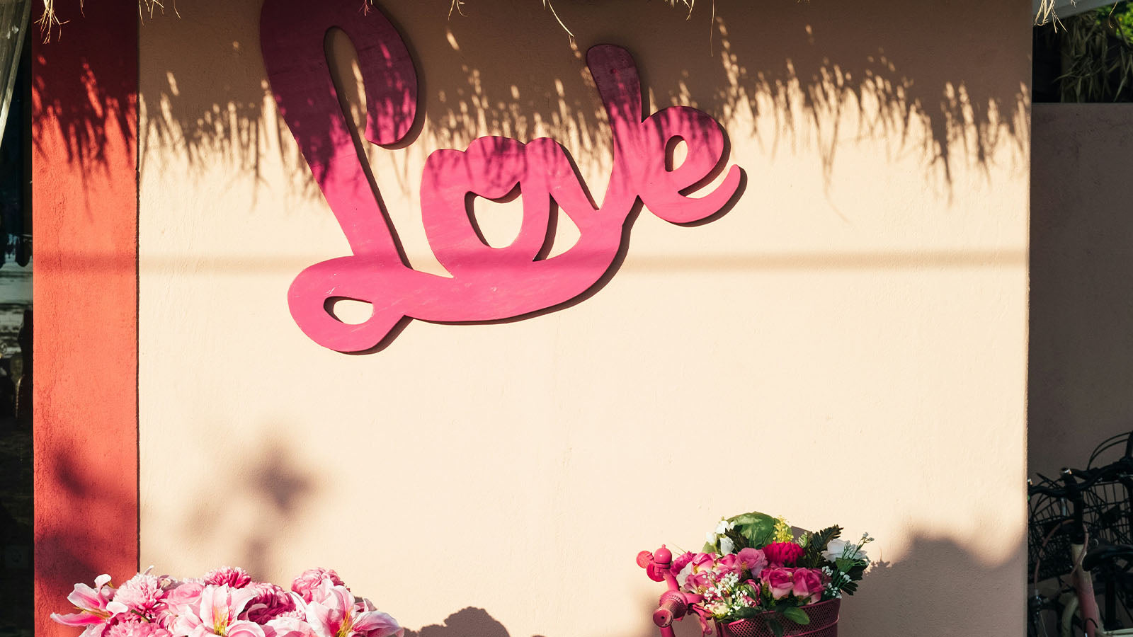 A large pink cut-out sign reading "Love" mounted on a cream wall above flowers in bright sunlight.