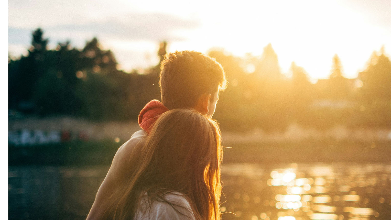 A couple seen from behind, standing together beside water at sunset.