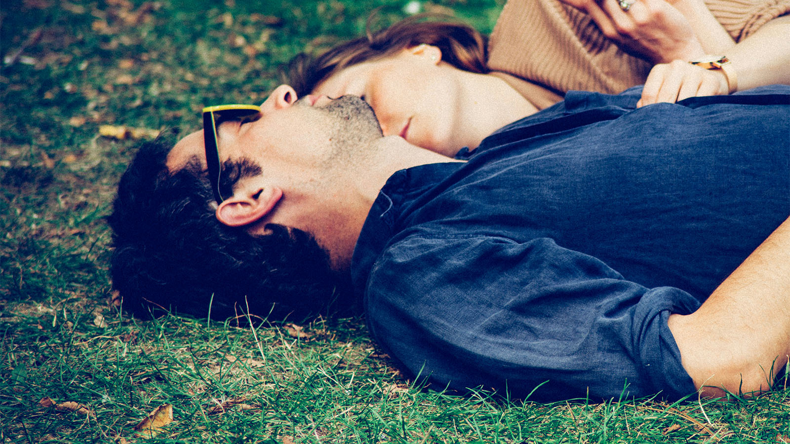 A couple lying on grass with their heads together, the man wearing sunglasses, seen from above.
