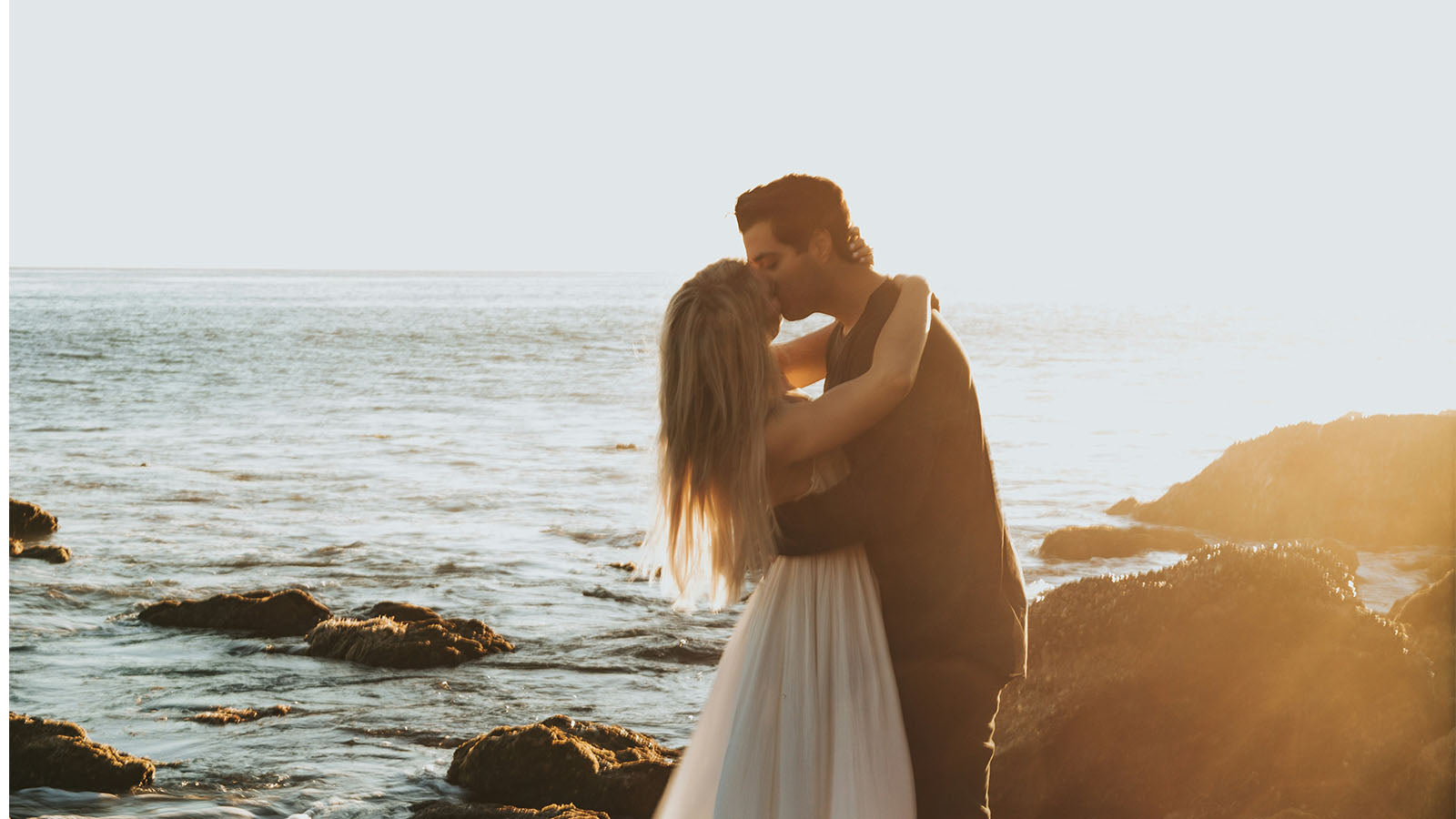 A couple embracing on rocks at the water's edge at sunset.