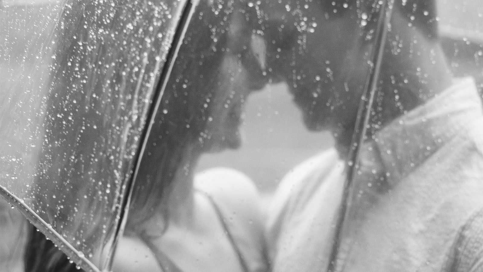 A black and white photograph of a couple standing close together beneath a rain-covered umbrella.