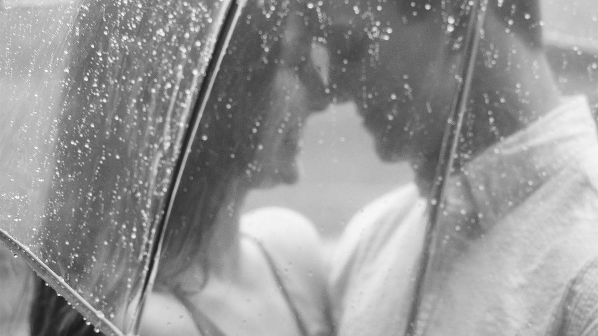 A black and white photograph of a couple standing close together beneath a rain-covered umbrella.