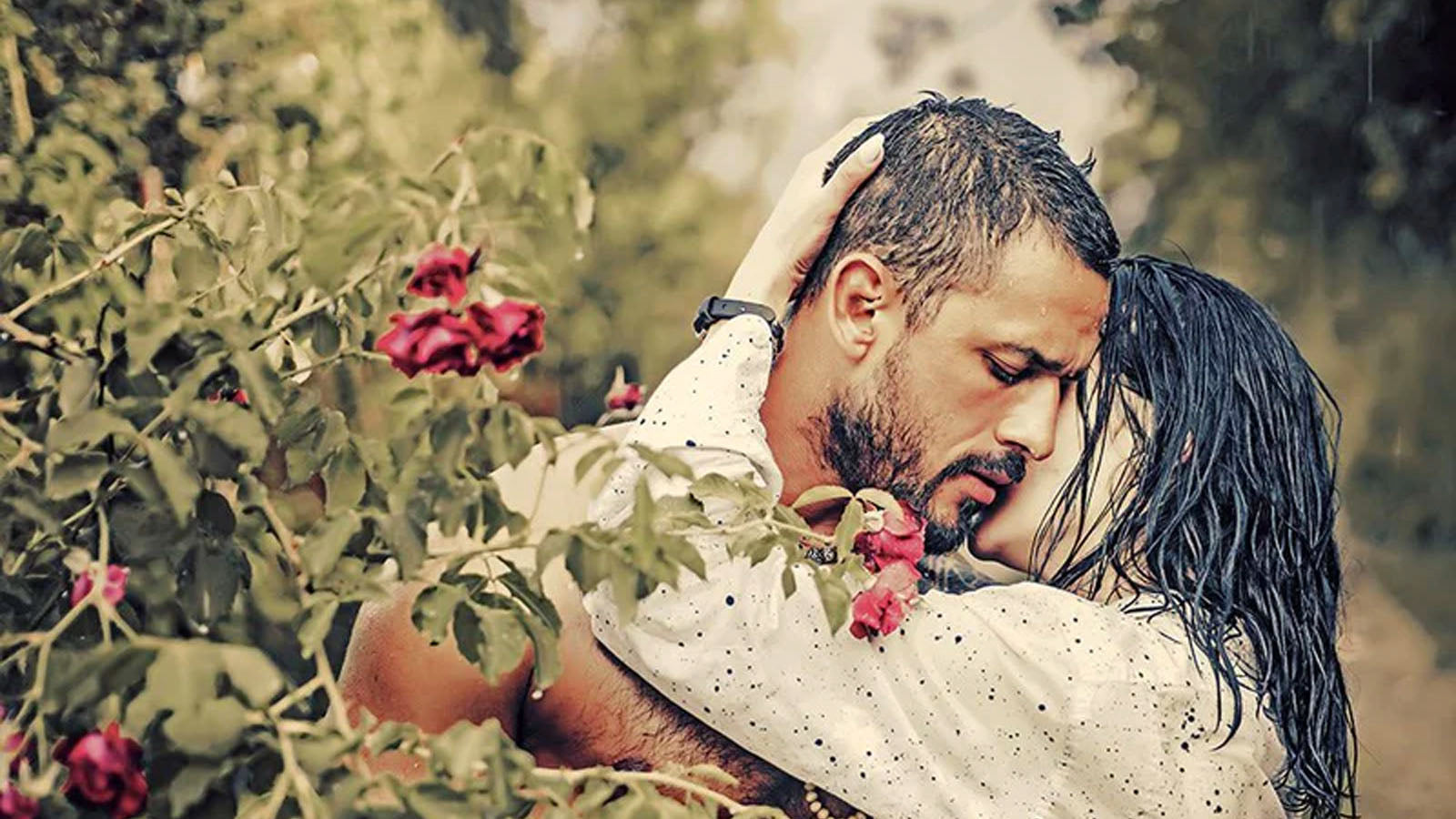 A couple embracing in the rain beside a rose bush, her hand resting on the back of his head.