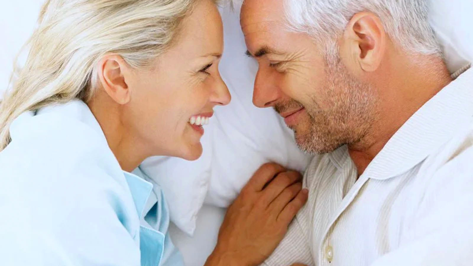 An older couple lying on white bedding, facing each other and smiling.