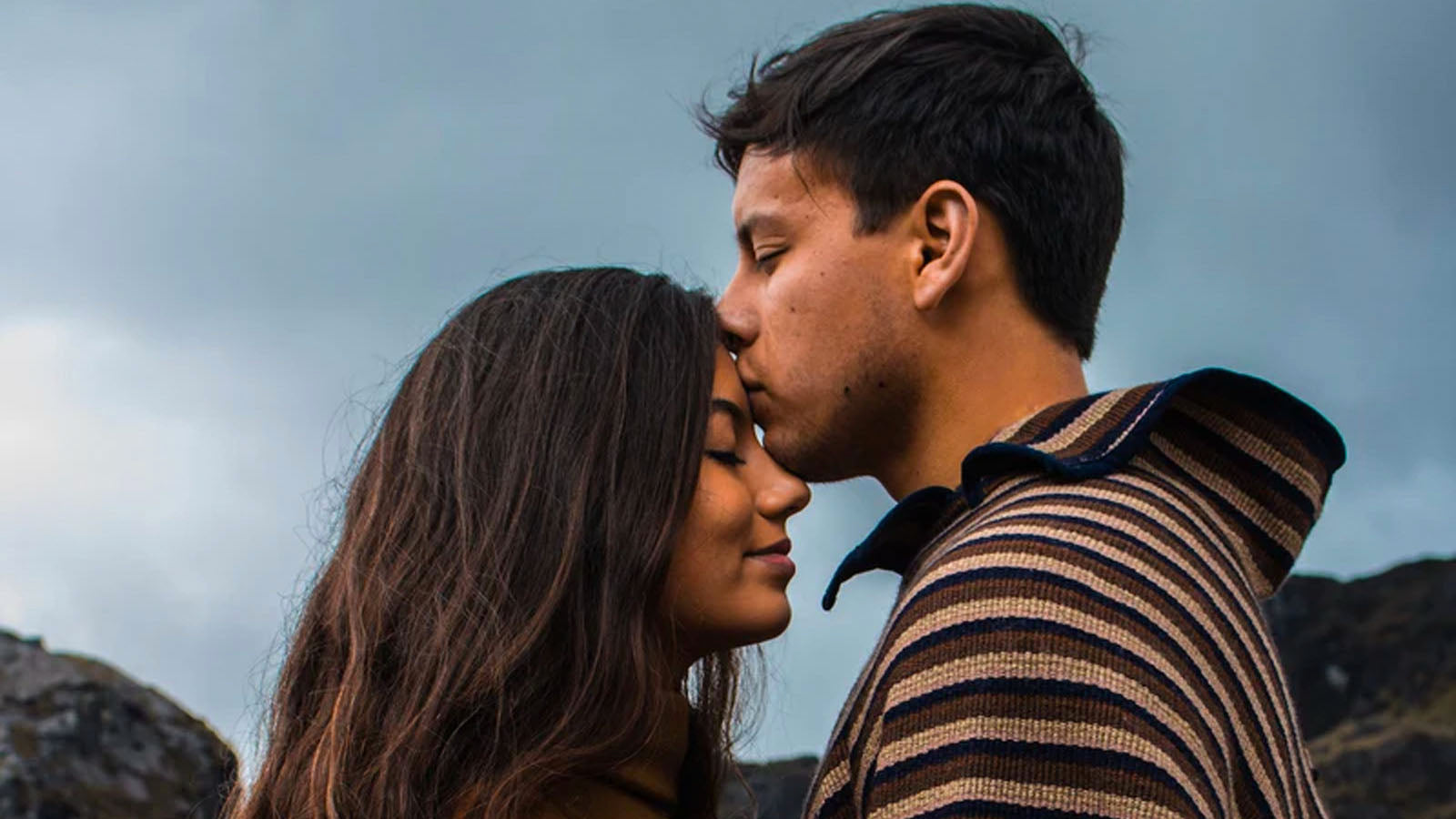A couple standing close together outdoors under a grey sky, his lips resting against her forehead.