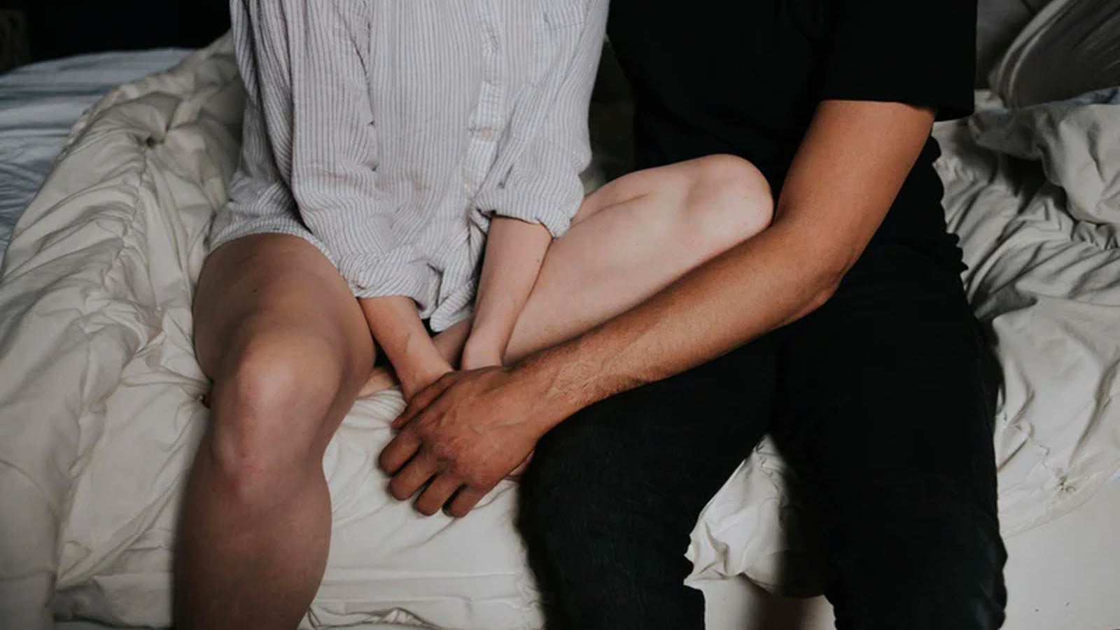 A couple sitting side by side on the edge of a bed, holding hands, shown from the waist down.