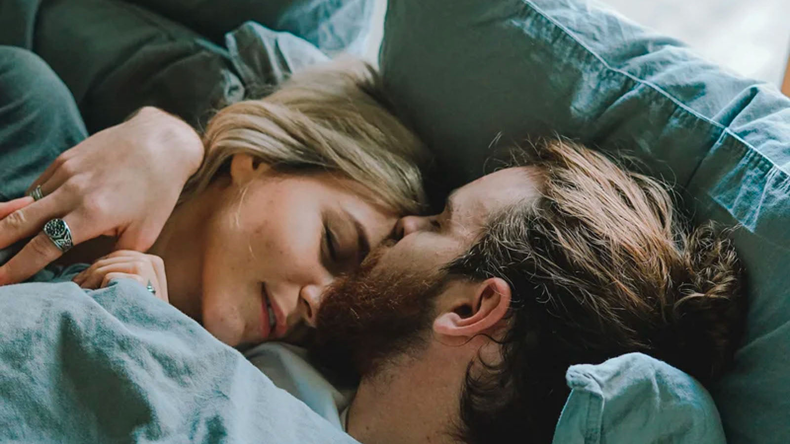 A couple lying close together in bed under green bedding, foreheads touching.