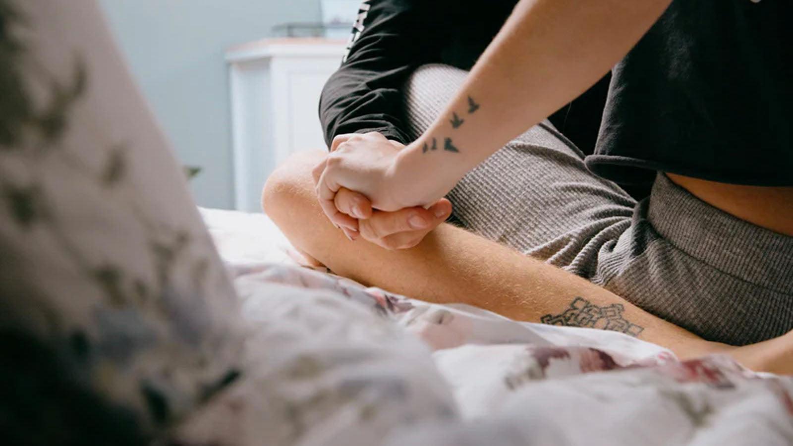 Two people sitting cross-legged on a bed, holding hands, shown from the waist down.
