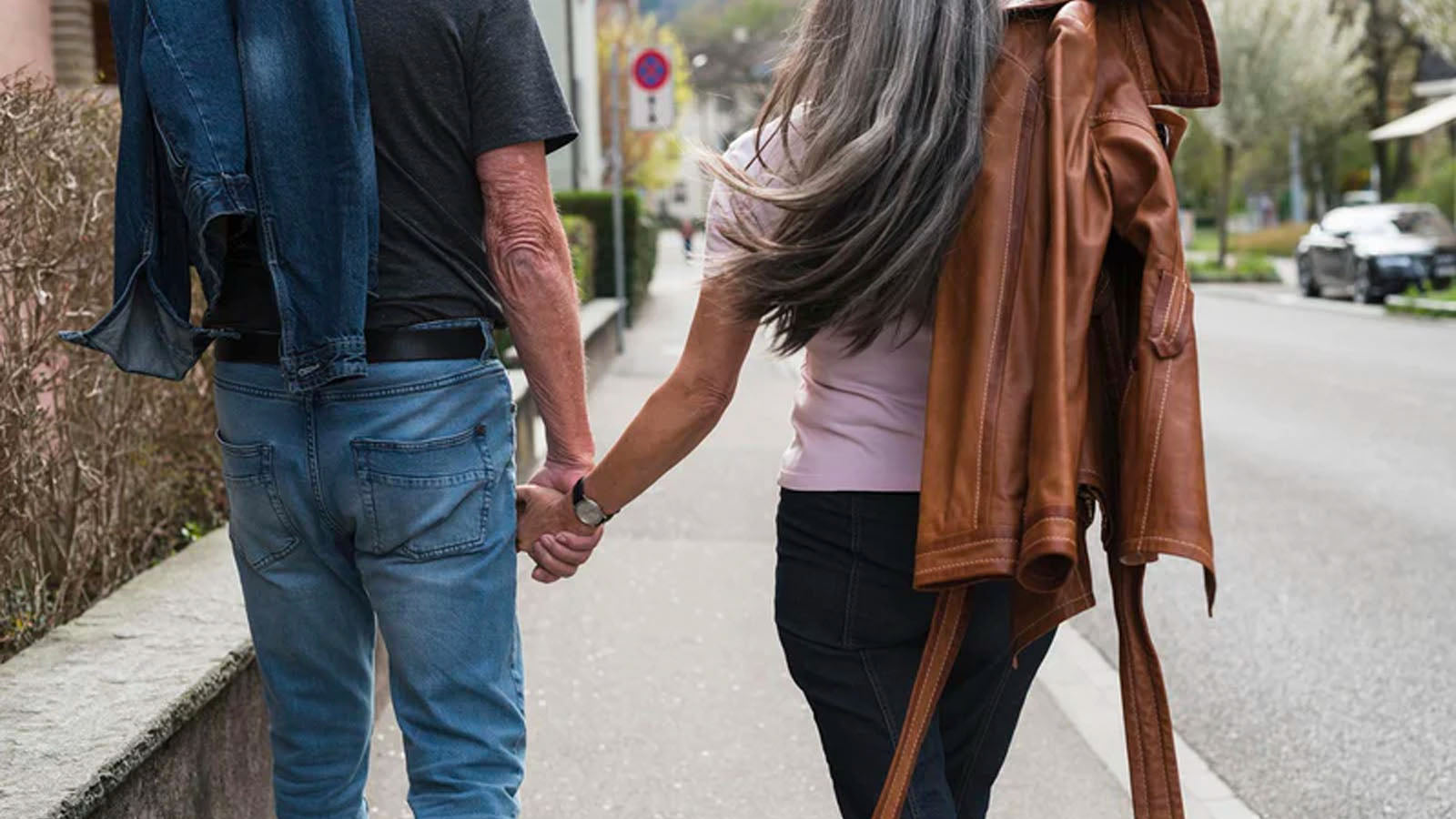 An older couple seen from behind, walking hand in hand along a suburban street.