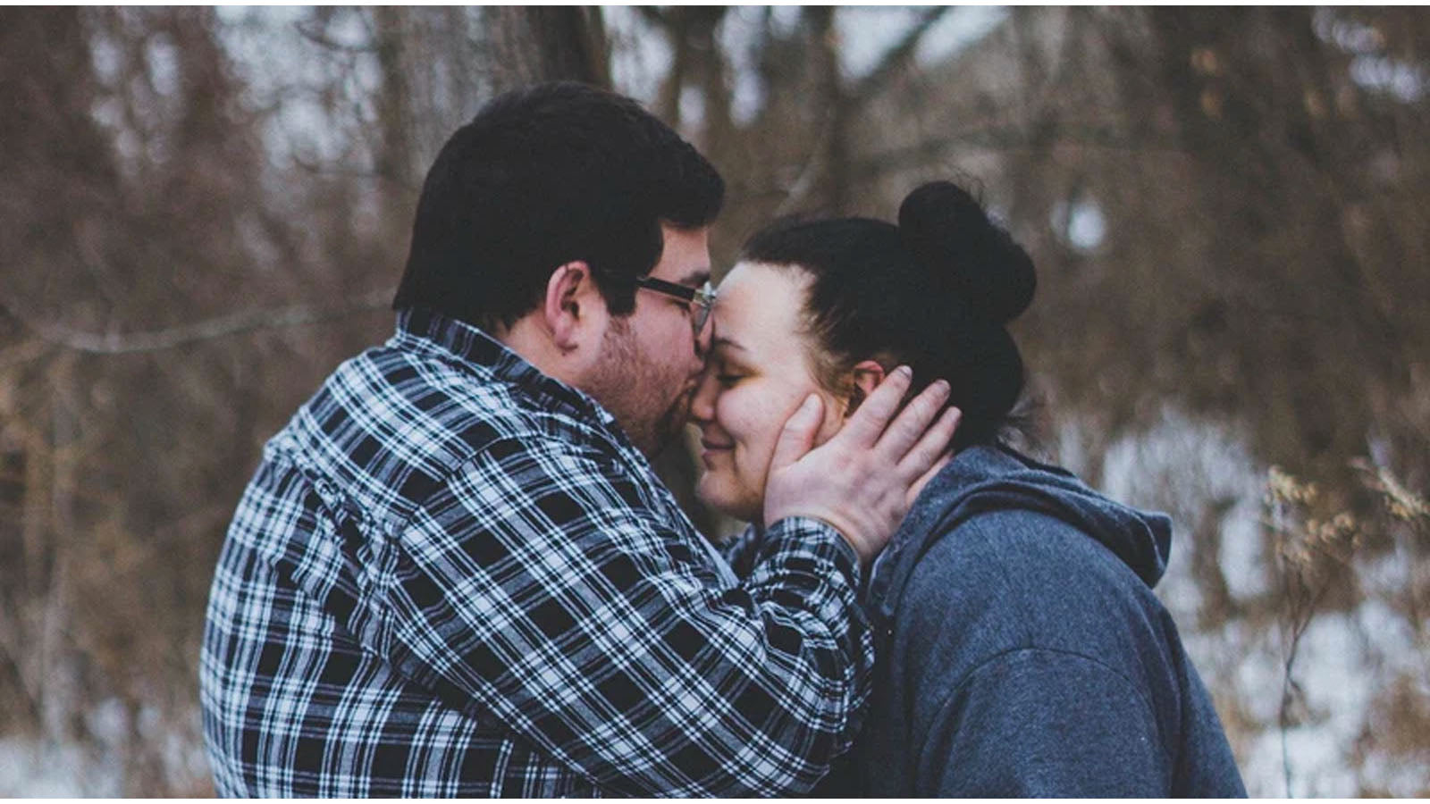 A couple embracing outdoors in winter, the man kissing the woman's forehead.