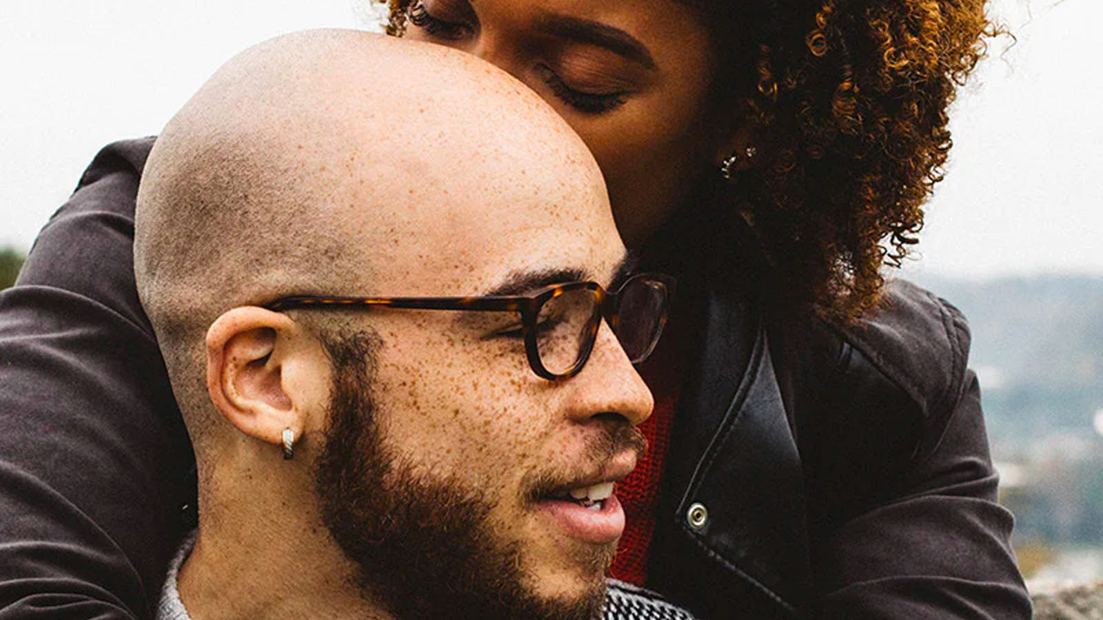 A close photograph of a couple outdoors, the woman resting her head against the man's shoulder from behind.