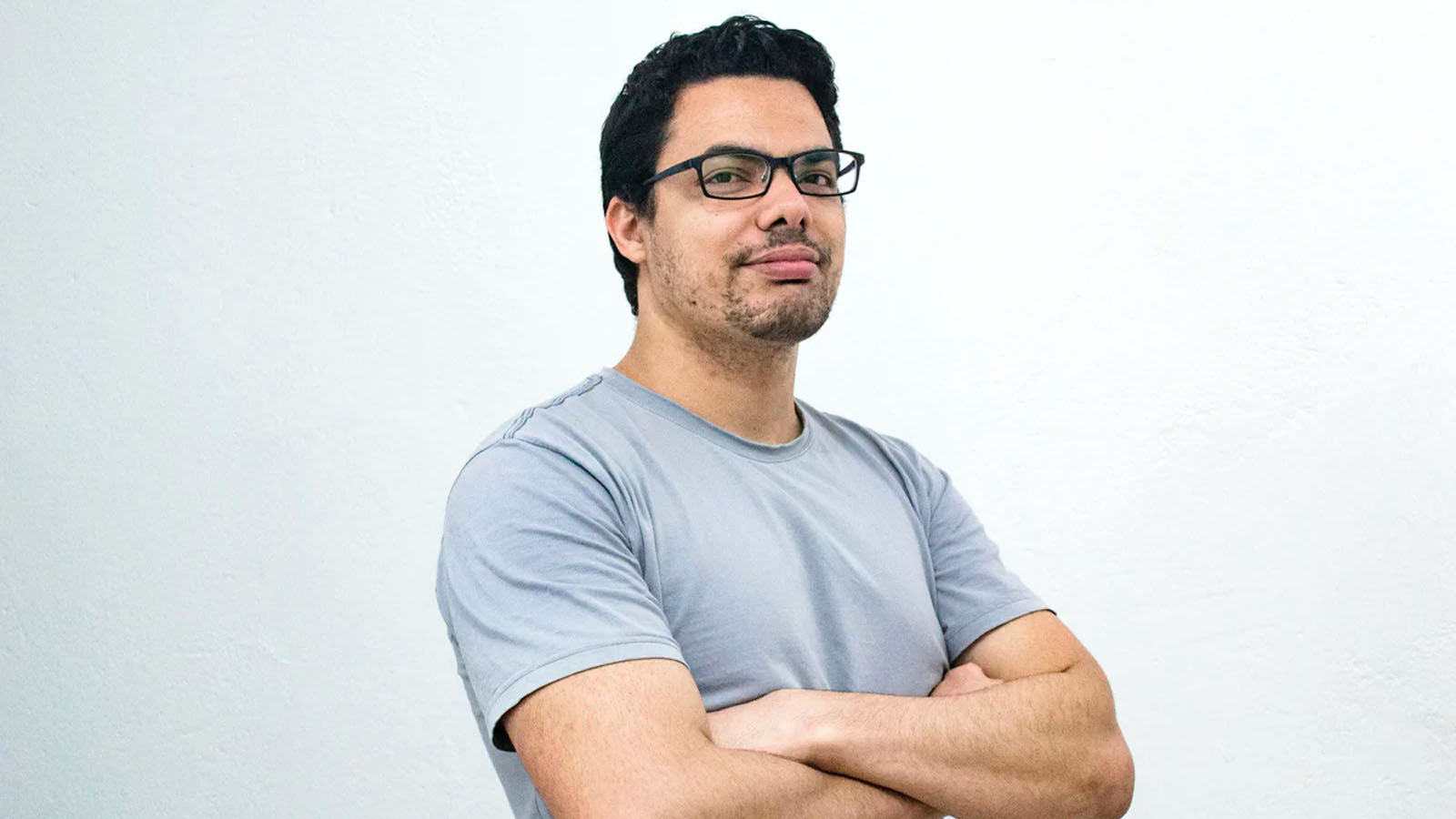A man in glasses with his arms folded, standing against a plain white wall.
