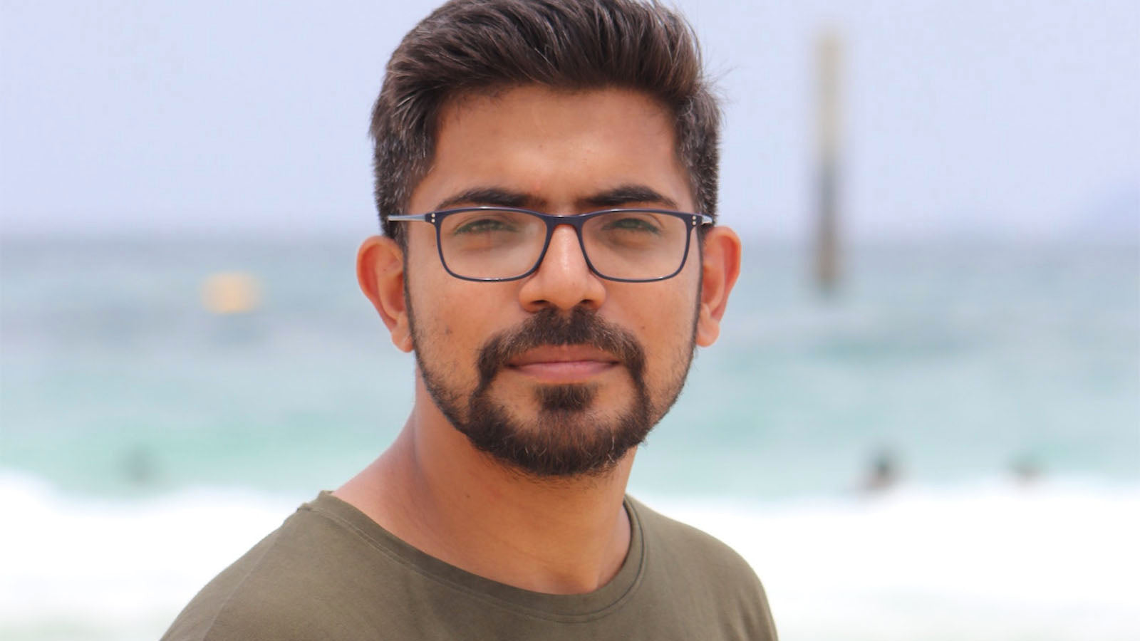 A man in glasses and an olive t-shirt looking at the camera, with the sea behind him.