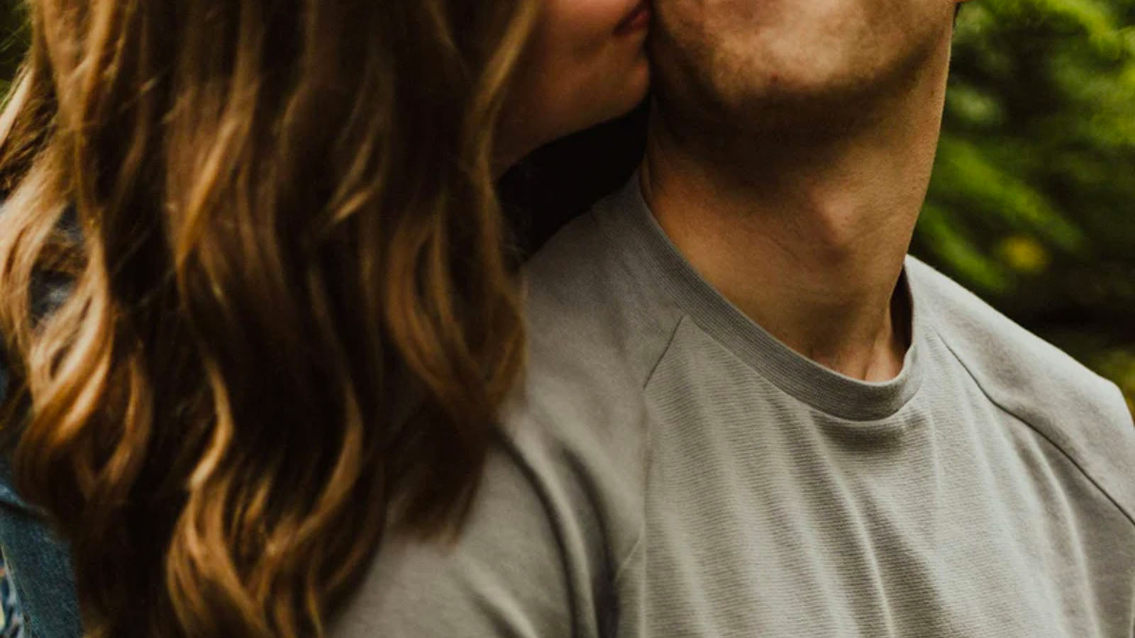 A close photograph of a woman kissing a man's neck, both shown from the shoulders up.