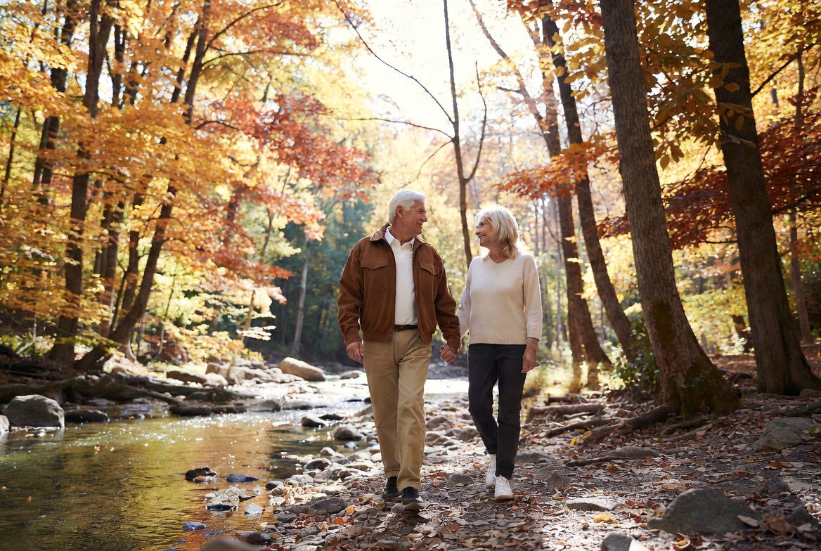 An older couple walking hand in hand beside a river through autumn woodland.